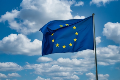 [ai] The European Union flag waving on a pole against a backdrop of blue sky and white clouds, featuring a blue field with a circle of twelve yellow stars.